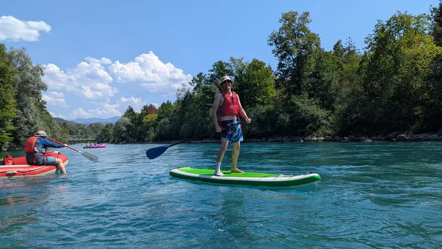 Mann steht auf einem Stand-up-Paddleboard auf der Aare, im Hintergrund ein rotes Schlauchboot