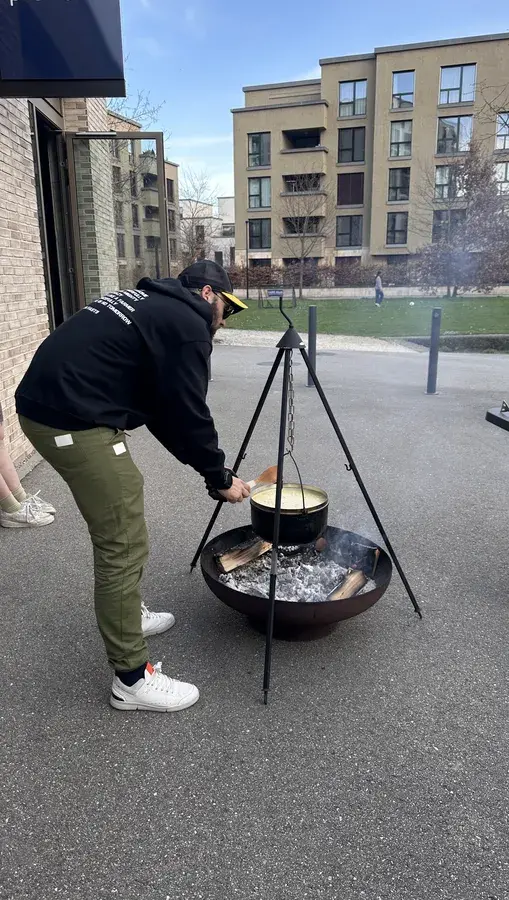 Eine Person in einem schwarzen Kapuzenpulli kocht draußen auf einem Dreifußgrill, im Hintergrund sind moderne Gebäude zu sehen.