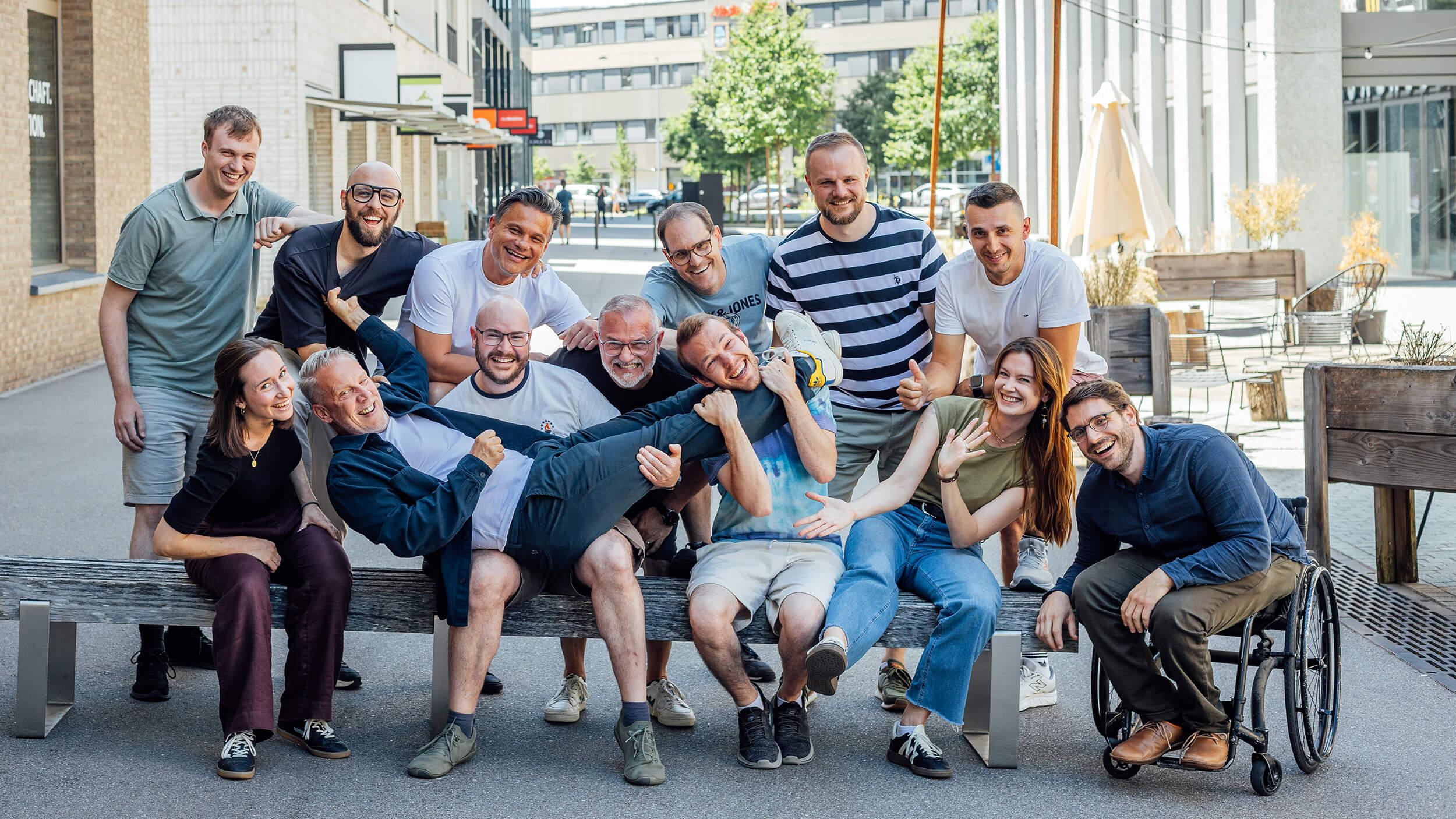 Eine Gruppe von dreizehn Menschen, die im Freien lachen und spielerisch posieren, wobei eine Person quer über ihren Schoss auf einer Bank liegt, was eine fröhliche Atmosphäre schafft.