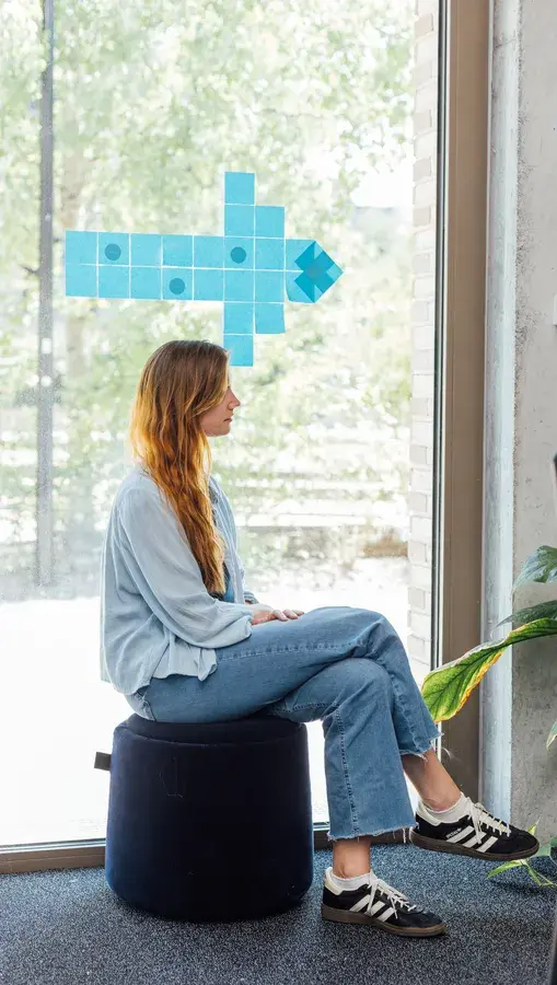 Eine Person mit langen Haaren, die ein hellblaues Outfit trägt, sitzt auf einem Hocker am Fenster, an dem ein Pfeil aus blauen Quadraten nach rechts zeigt.