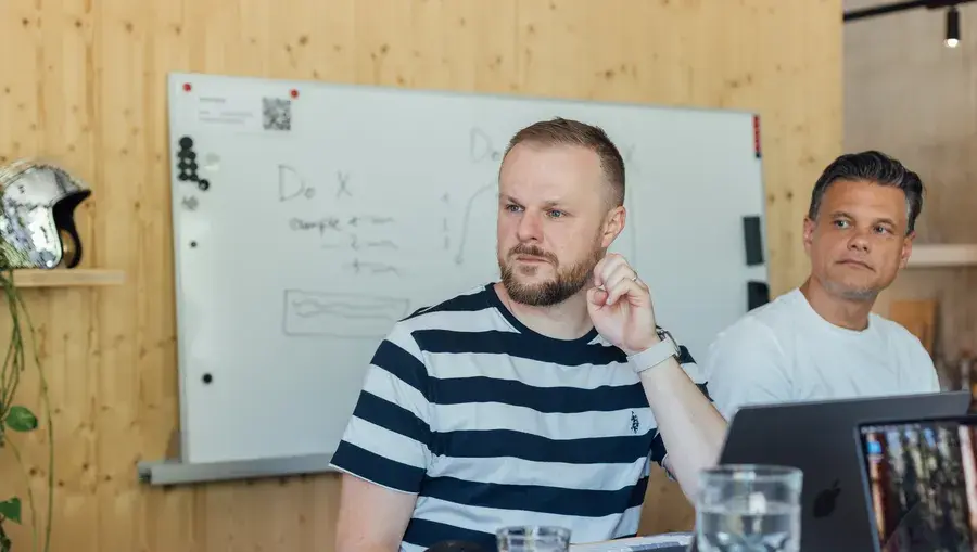 Zwei Männer sitzen an einem Tisch in einem Besprechungsraum; einer trägt ein gestreiftes Hemd. Auf dem Tisch stehen ein Laptop und Wassergläser, im Hintergrund ist ein Whiteboard zu sehen.