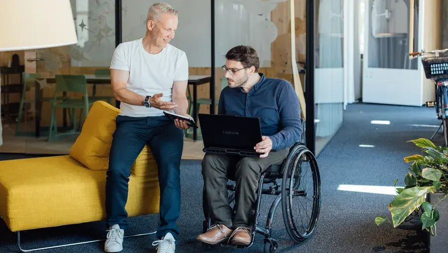 Zwei Männer in einem Büro: Der eine sitzt mit einem Laptop im Rollstuhl, während der andere, in Freizeitkleidung, sich mit ihm unterhält.