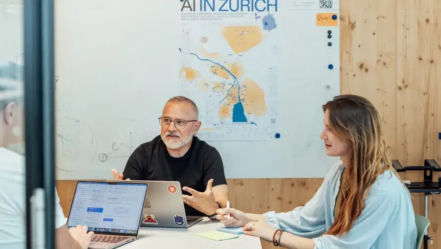 Drei Mitarbeitende sitzen im Gespräch an einem Tisch mit Laptops