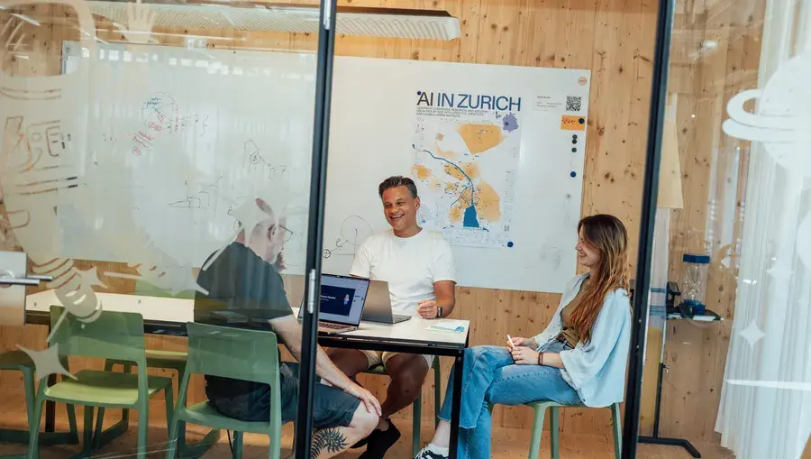 Drei Personen halten in einem modernen Konferenzraum mit Holzausstattung eine Besprechung ab. Auf einem Schild an der Wand steht „KI in Zürich“.