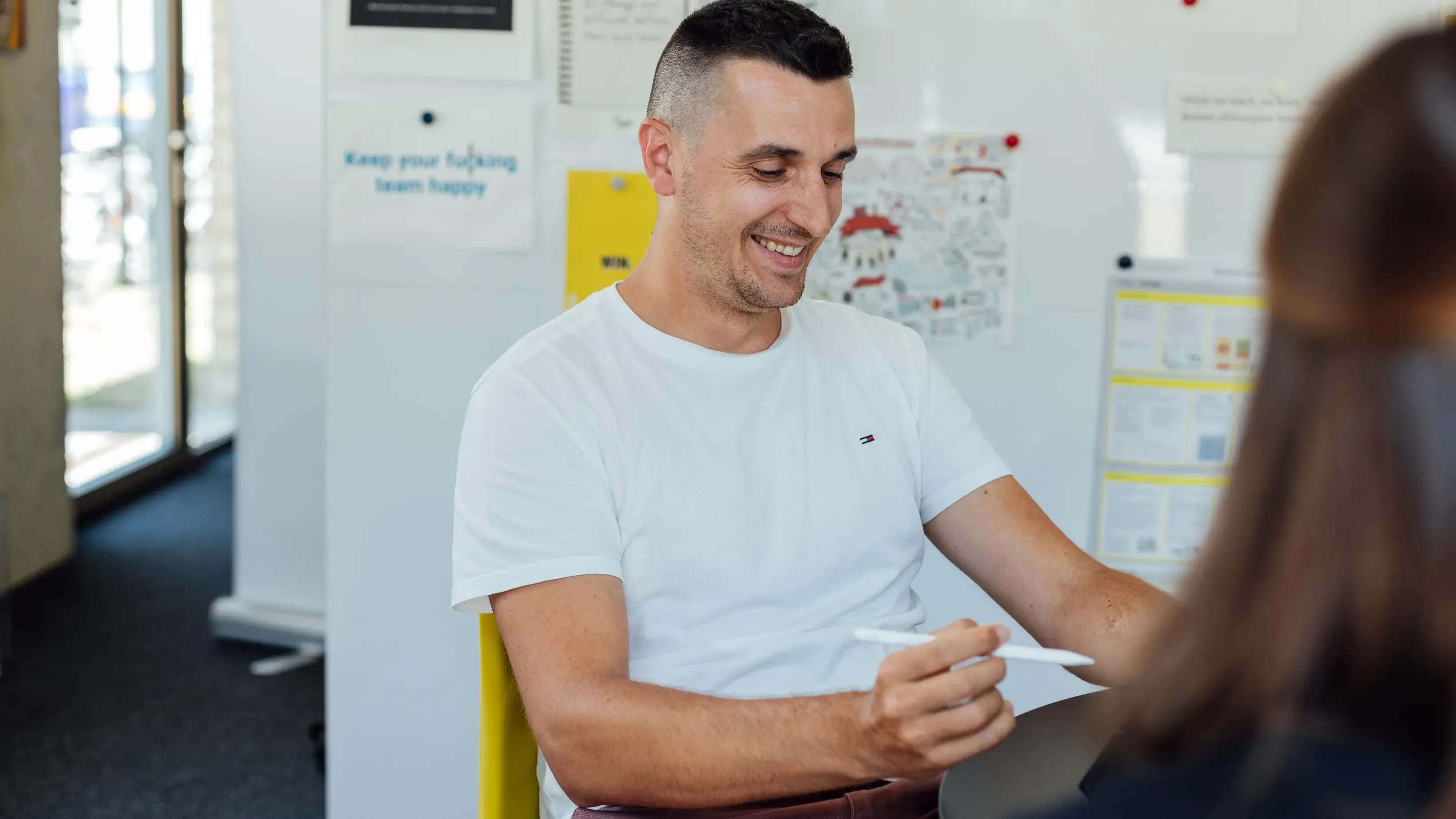 Ein Mann in einem weißen T-Shirt sitzt lächelnd an einem Tisch und hält Stift und Papier in der Hand. Im Hintergrund ist ein Büro mit Plakaten zu sehen, und durch eine Glastür fällt Licht herein.
