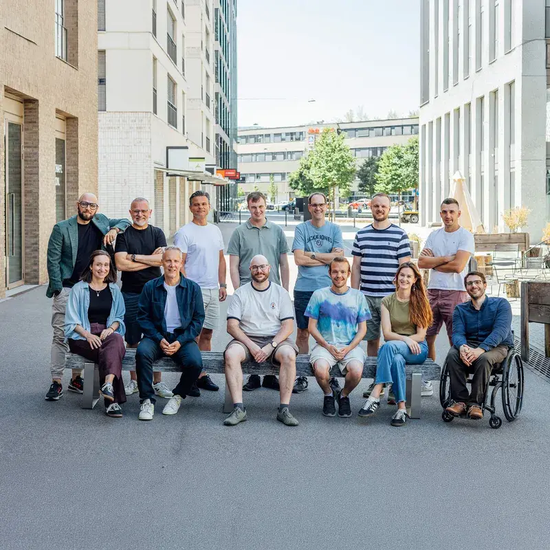 Eine Gruppe von dreizehn Personen, darunter eine im Rollstuhl, posiert gemeinsam im Freien vor einer modernen städtischen Kulisse, lächelnd und leger gekleidet.