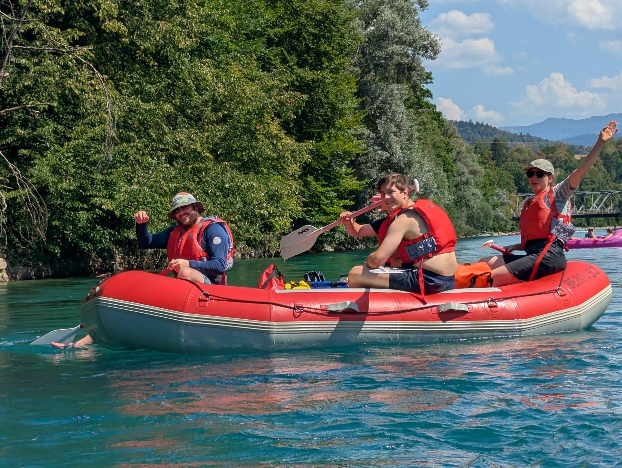 Drei Personen mit Schwimmwesten winken fröhlich aus einem roten Schlauchboot auf der Aare