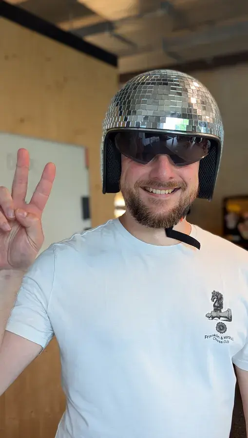 Ein lächelnder Mann, der einen glänzenden Helm in Form einer Discokugel und eine Sonnenbrille trägt, mit der rechten Hand das Peace-Zeichen macht und in einem Innenraum steht.