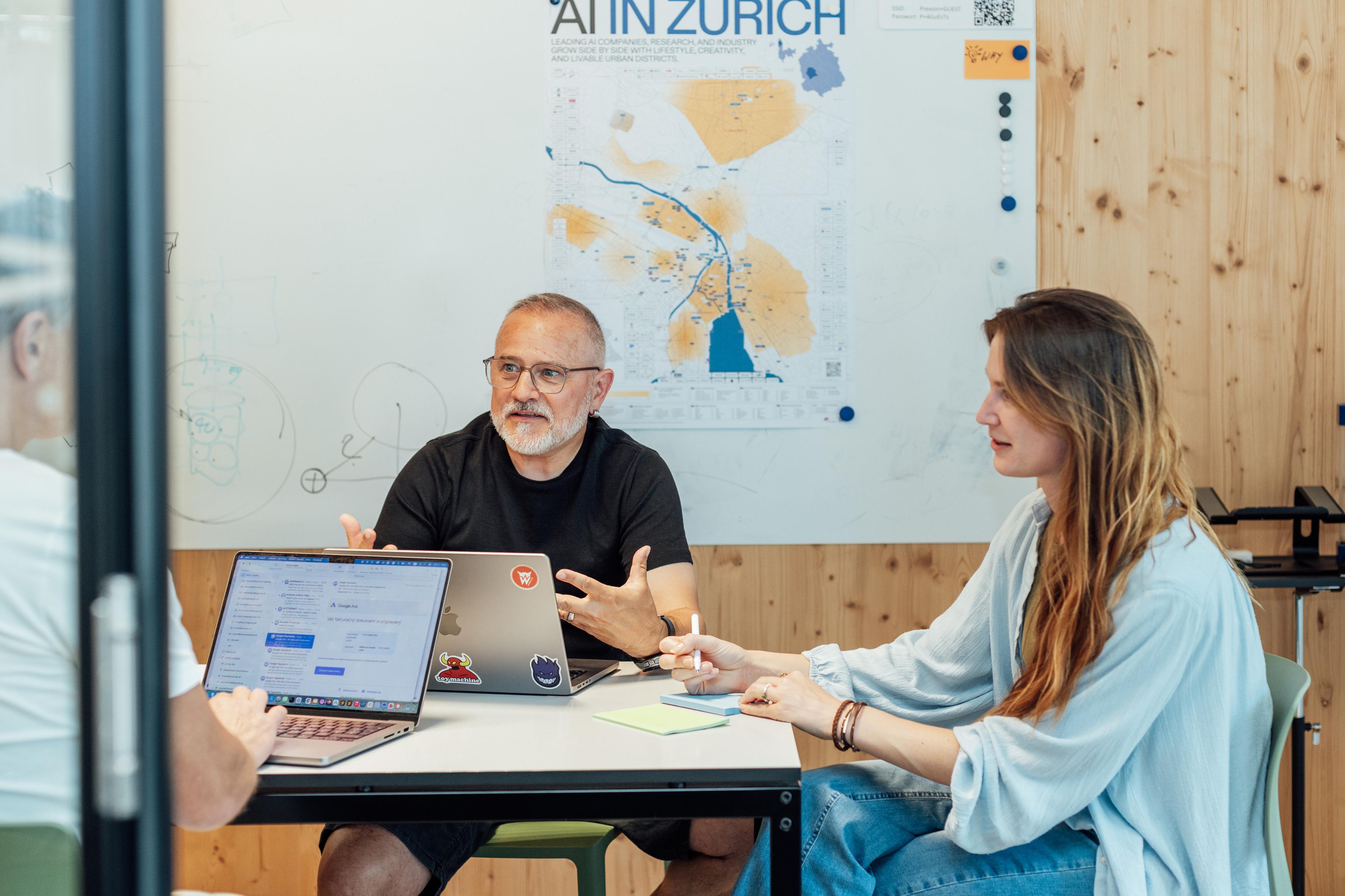 Drei Personen sitzen in einem Besprechungsraum und unterhalten sich. Ein Mann spricht, während die beiden anderen zuhören; ihre Laptops liegen offen auf dem Tisch.