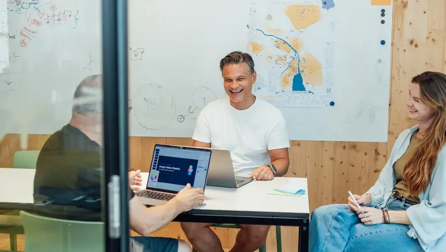 Drei Personen sitzen in einem Besprechungsraum um einen Tisch herum und diskutieren, während sie Laptops und Unterlagen vor sich haben. An der Wand hinter ihnen hängen eine Karte und Notizen.